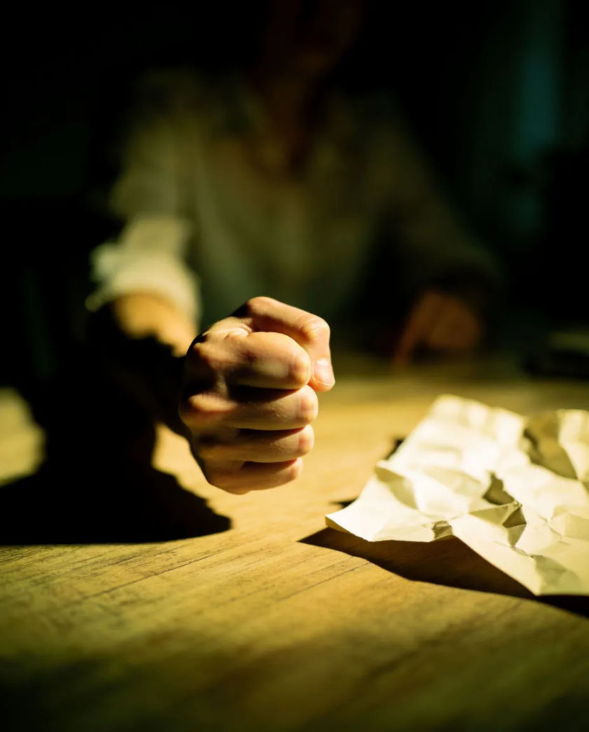 A clenched fist trembling above a plain wooden table, the knuckles pale, a crumpled sheet of paper beside it under harsh overhead light
