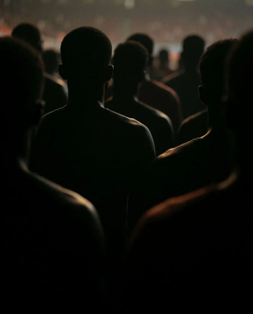 A crowd of dark silhouettes standing close together, faces turned the same way, soft warm light catching a few shoulders