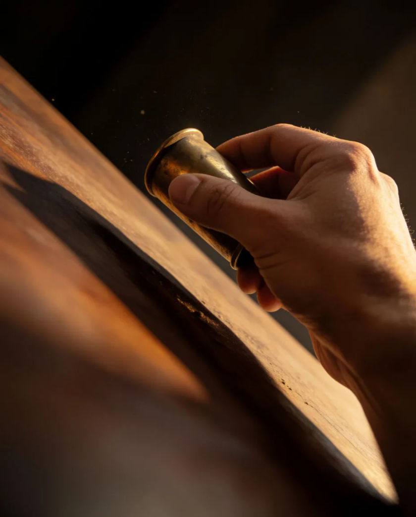 A hand closing around a small brass object lifted just off a dark wooden surface, a thin line of light catching its edge against a black ground