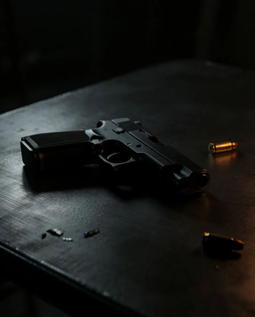 A plain matte black semi-automatic handgun silhouette resting on a dark steel table, a single brass casing catching a thin line of amber light beside it