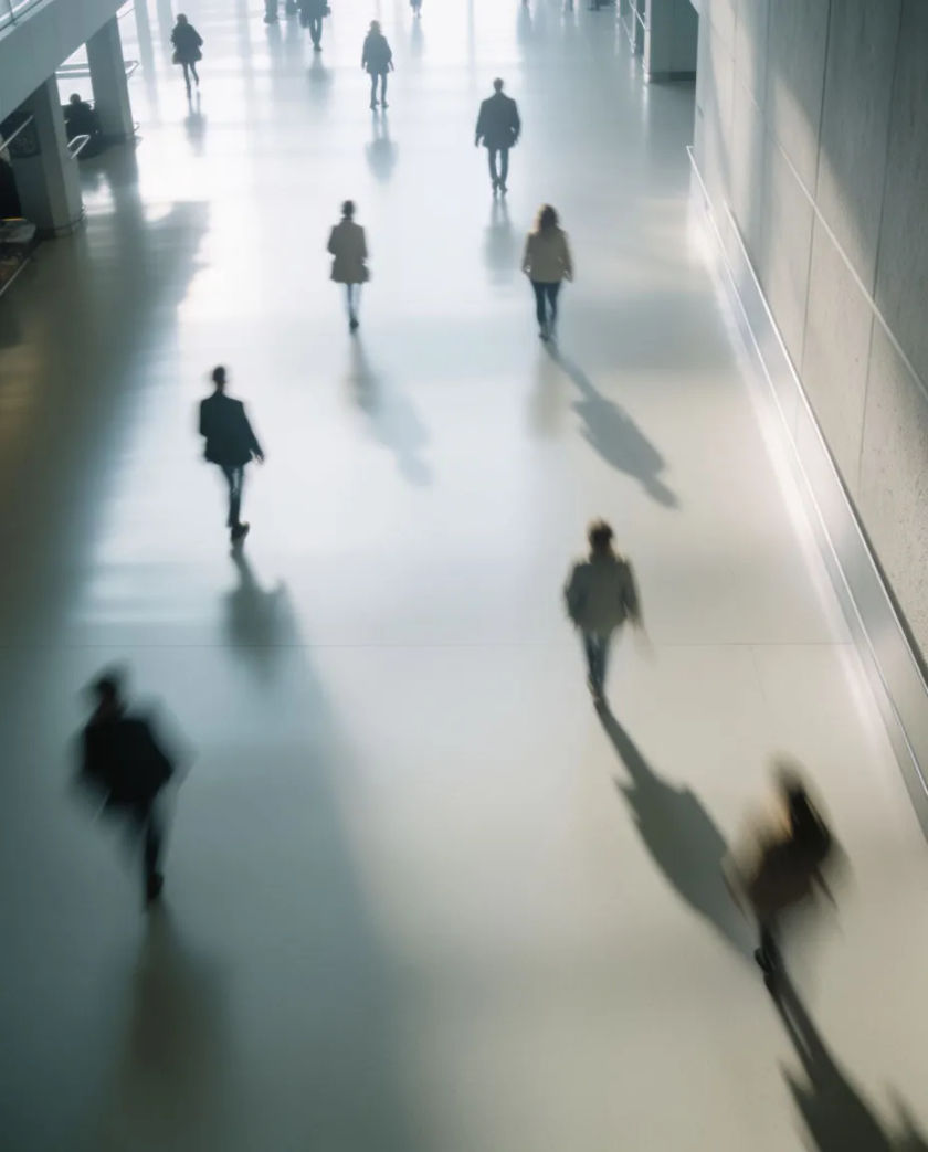 A wide pale concourse seen from above with small blurred figures crossing in different directions, long soft shadows on a polished floor
