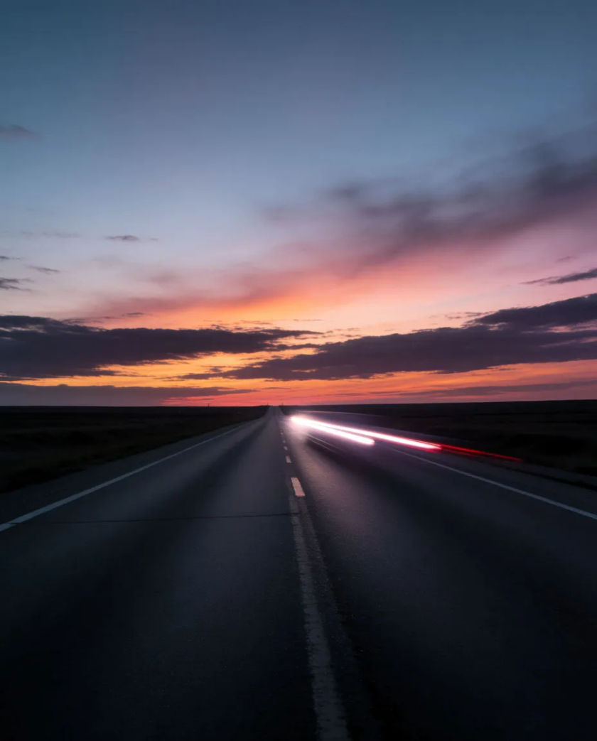 A long empty asphalt road narrowing toward a horizon, a single pair of headlight streaks smeared across the frame in the dusk