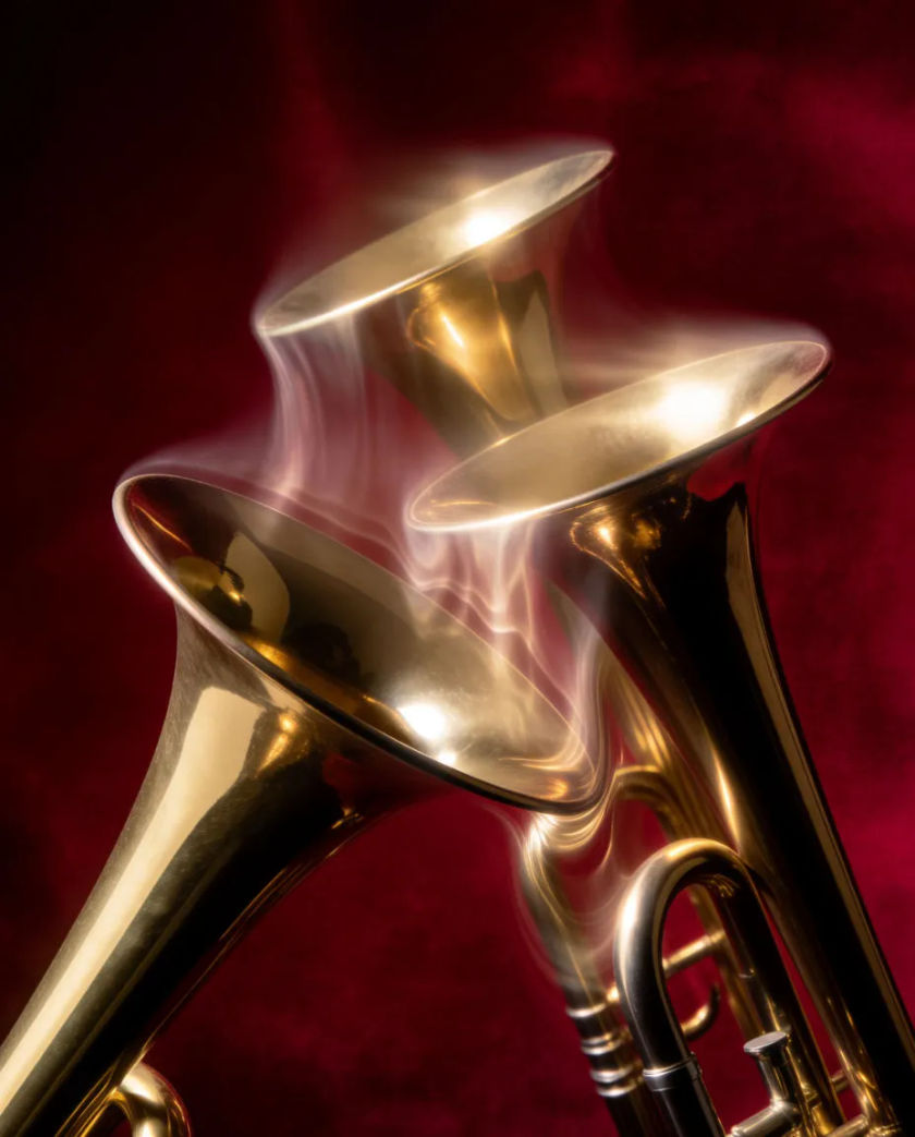 Three long brass trumpets raised together at an angle, gold bells catching light against a dark red velvet drape