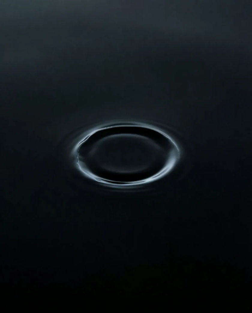 A single thin ripple of pale light spreading outward across a perfectly still black surface, the rest of the frame empty and dark
