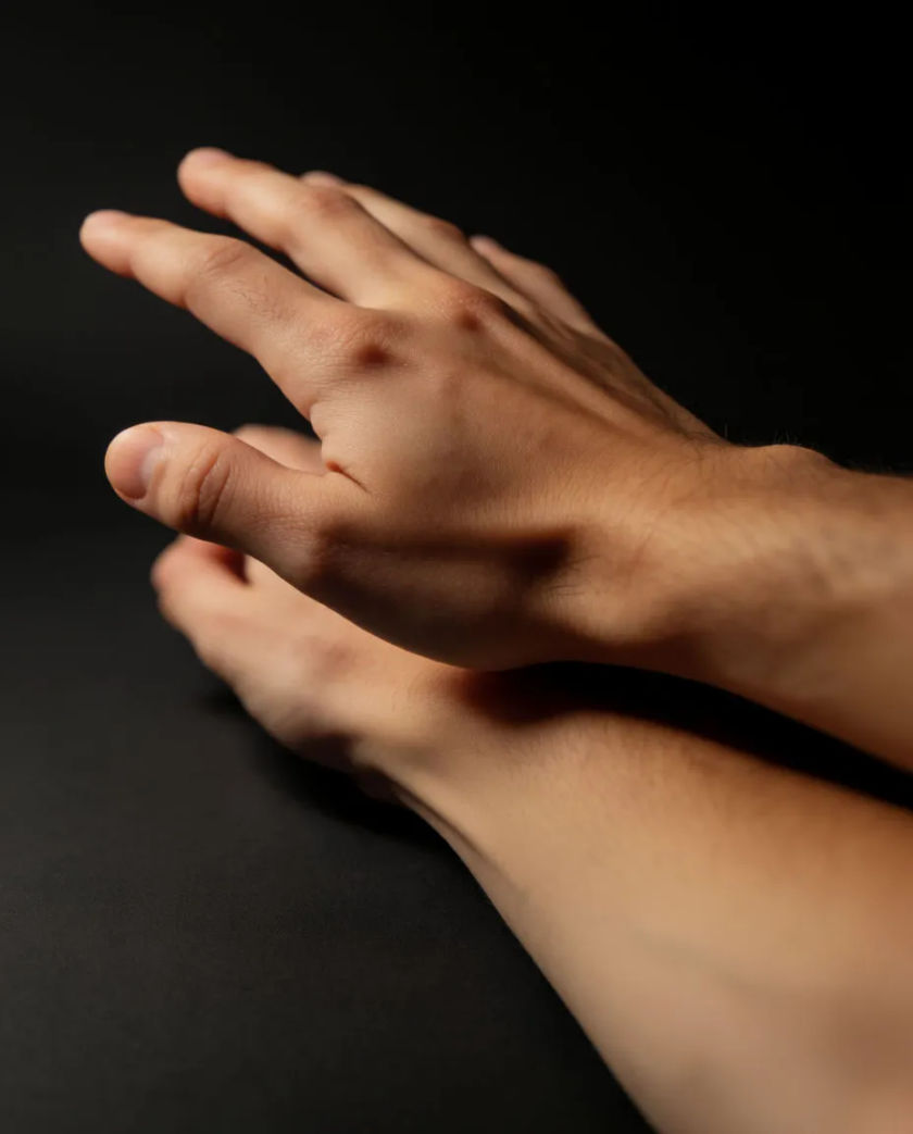 A bare forearm resting on a matte black surface, a hand hovering just above it in soft side light, fine skin texture visible