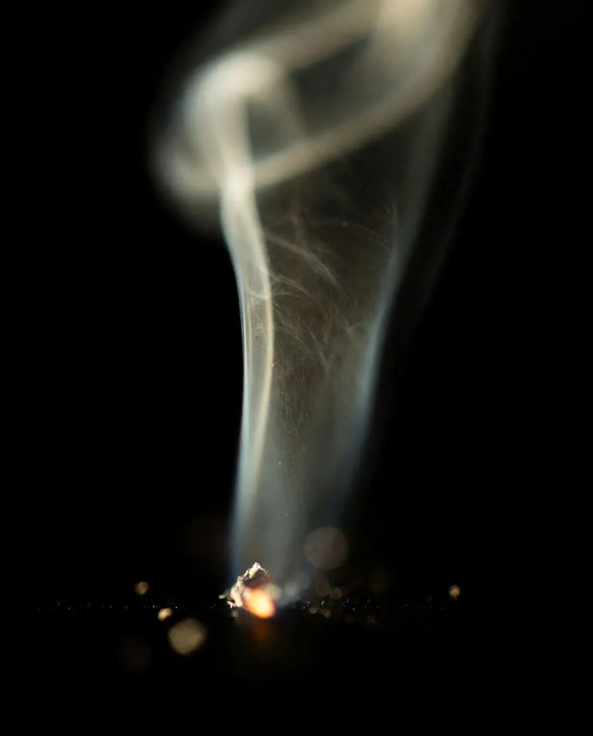 A thin ribbon of pale smoke curling up from a glowing ember against a pure black background, the ember lighting a few grains of ash