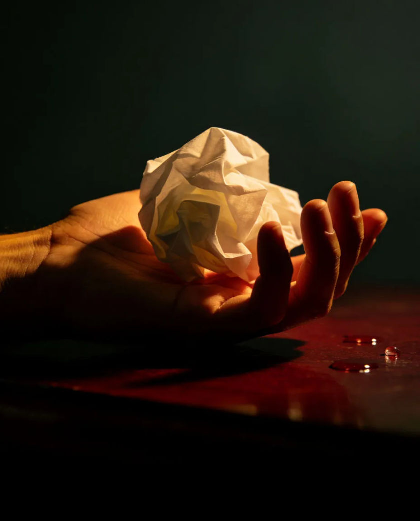 A single tissue crumpled in a hand resting on a dark table, a soft pool of cold light on the knuckles and a few small drops on the wood