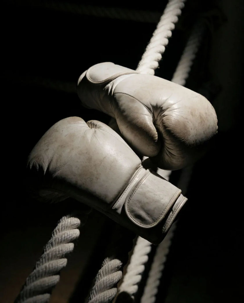 A pair of worn red boxing gloves hanging on a hook against a dark wall, a single hard light catching the cracked leather