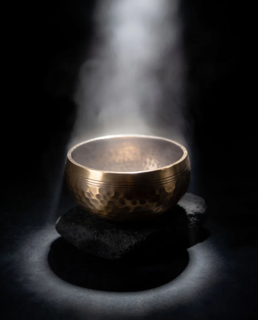 A single shaft of soft white light falling on a bronze singing bowl resting on dark stone, a fine haze rising around it
