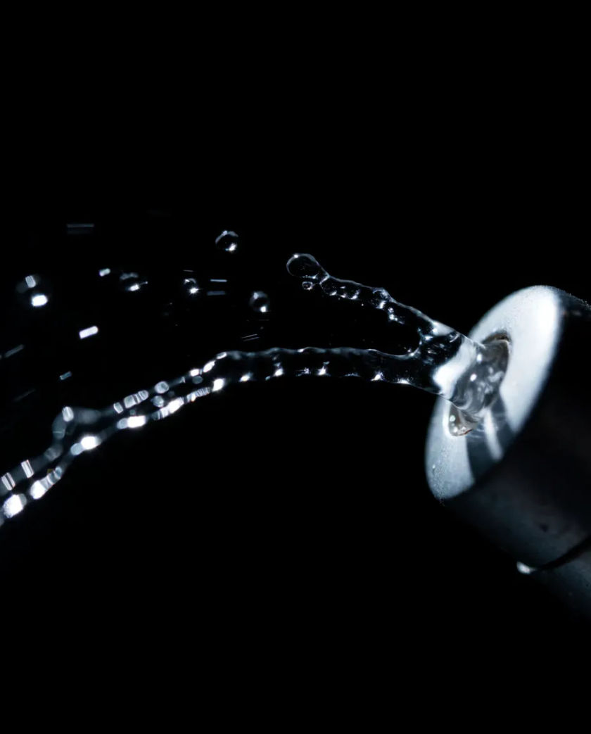 A thin arc of clear liquid leaping from a small nozzle against a black backdrop, each droplet caught in sharp white light