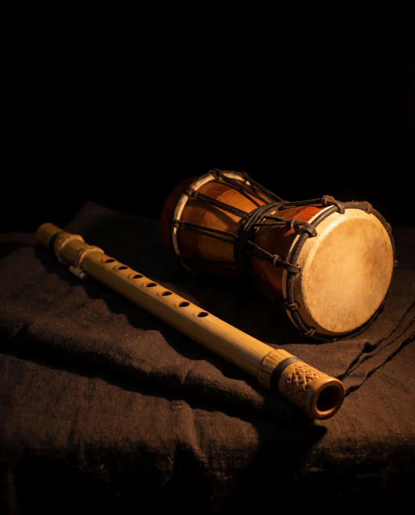 A bamboo flute and a small two-headed hand drum resting on a dark cloth, lit by one warm lamp against a black background