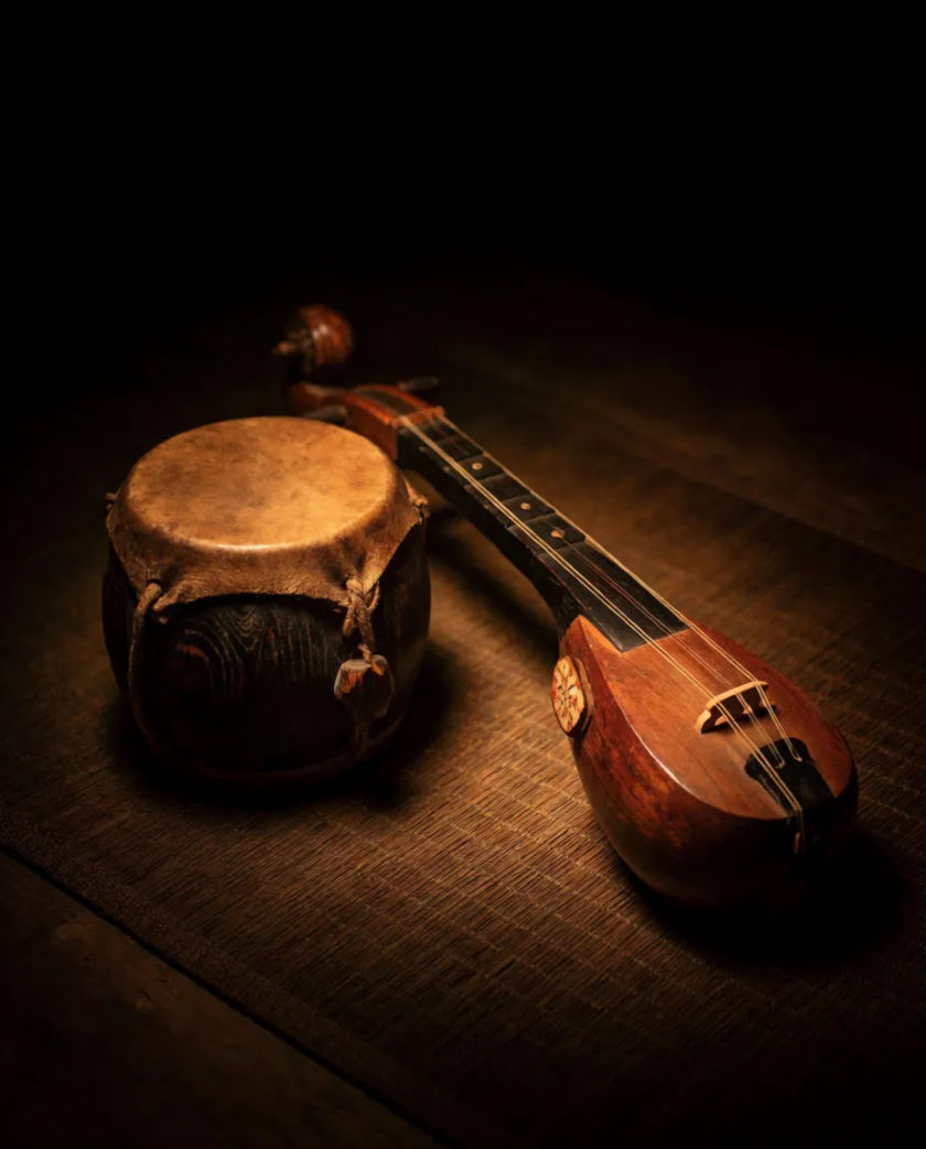 A small barrel-shaped drum and a plucked string instrument laid on a dark woven mat, a single warm lamp glowing against a black background