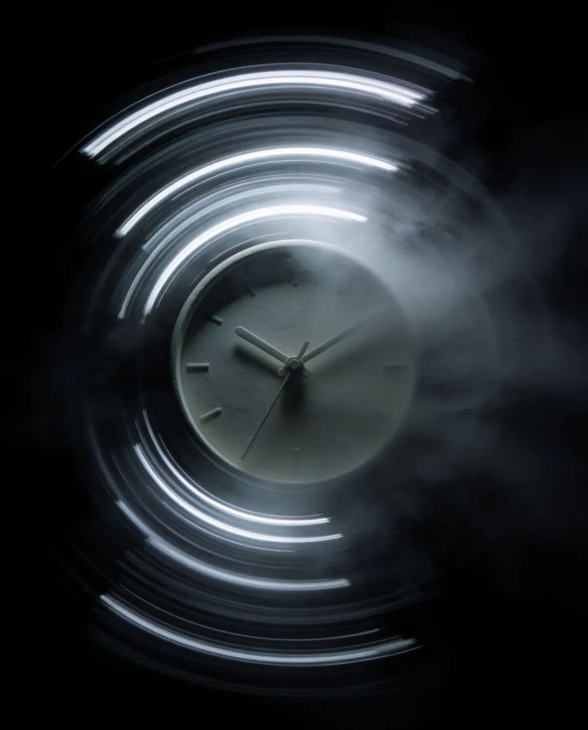 A thin ring of pale light streaking into a blur of concentric arcs around a still clock face on a pure black ground