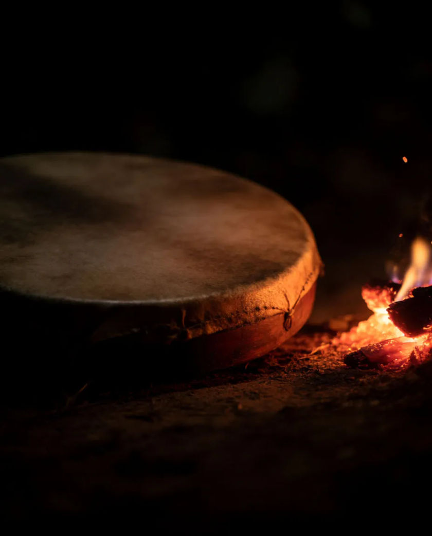 A large hide-covered hand drum on bare earth beside a glowing ember, sparks rising into a dark sky, warm texture on the drumskin