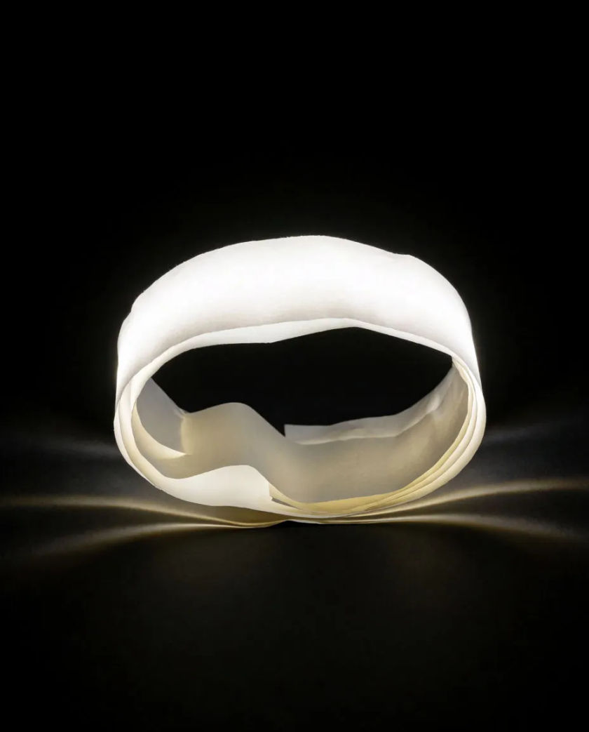 A plain glowing ring of soft white light unfolding on a matte black ground, thin ripples of brass-coloured light spreading outward from its centre