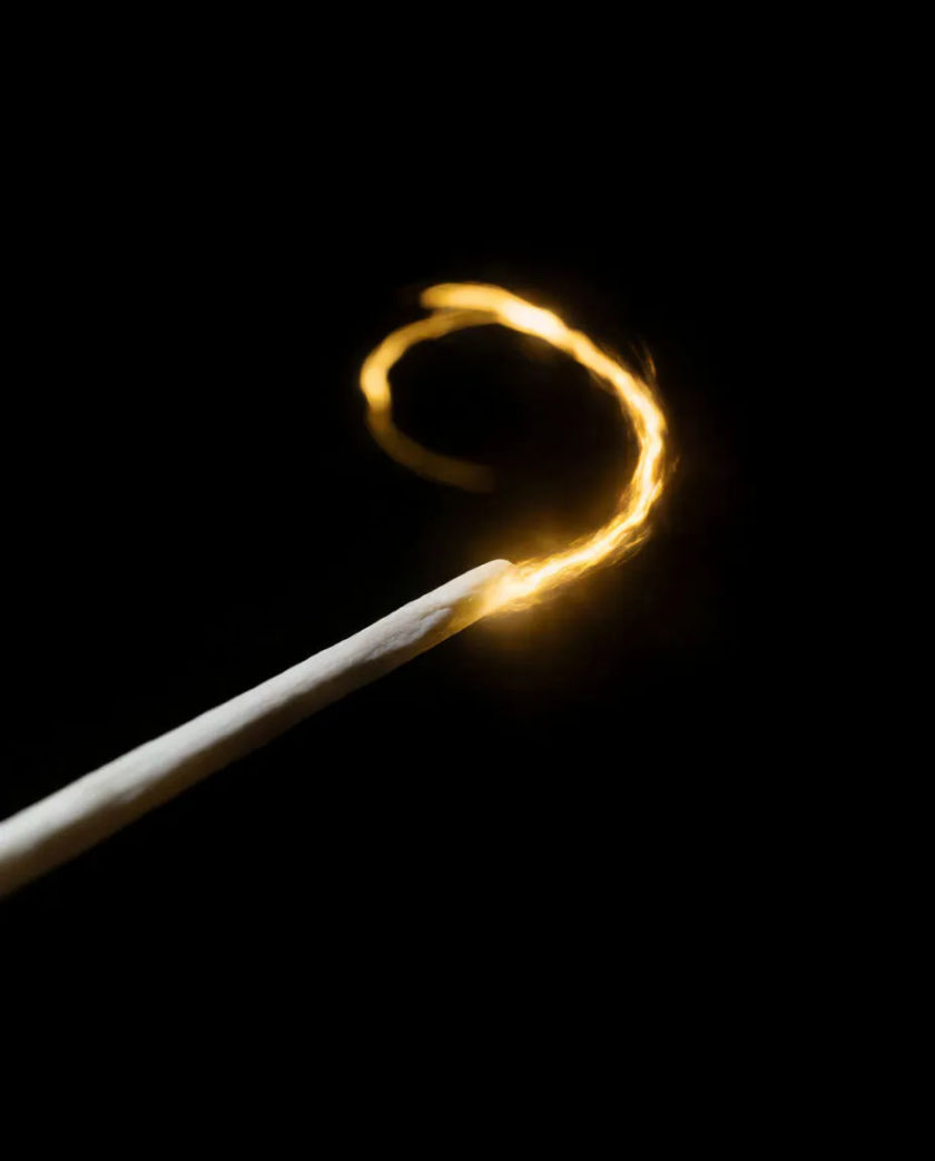 A thin pale wand held at an angle with a spiral of golden sparks trailing from its tip on a matte black ground, motion blur on the arc