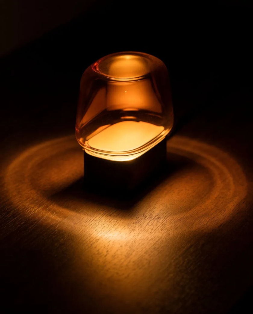 A glass lamp glowing amber on a dark wooden table, soft concentric halos spreading over the grain