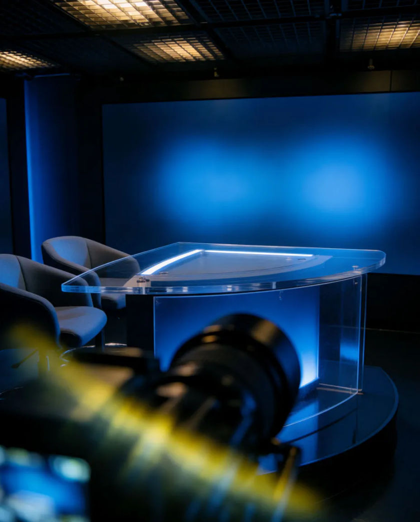 A dark television studio with a curved glass anchor desk lit by cool blue panels and a blank wall of screens behind it
