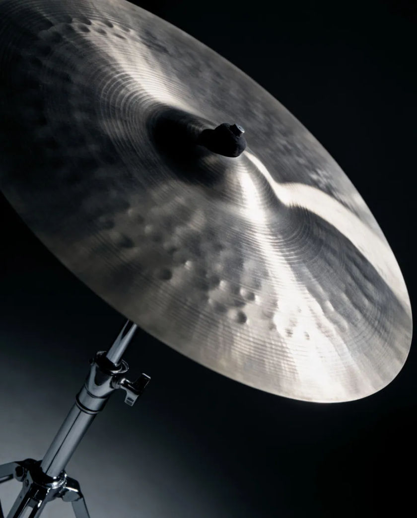 A large brass crash cymbal tilted on a stand with a sweep of pale light curving toward its edge against a dark studio backdrop