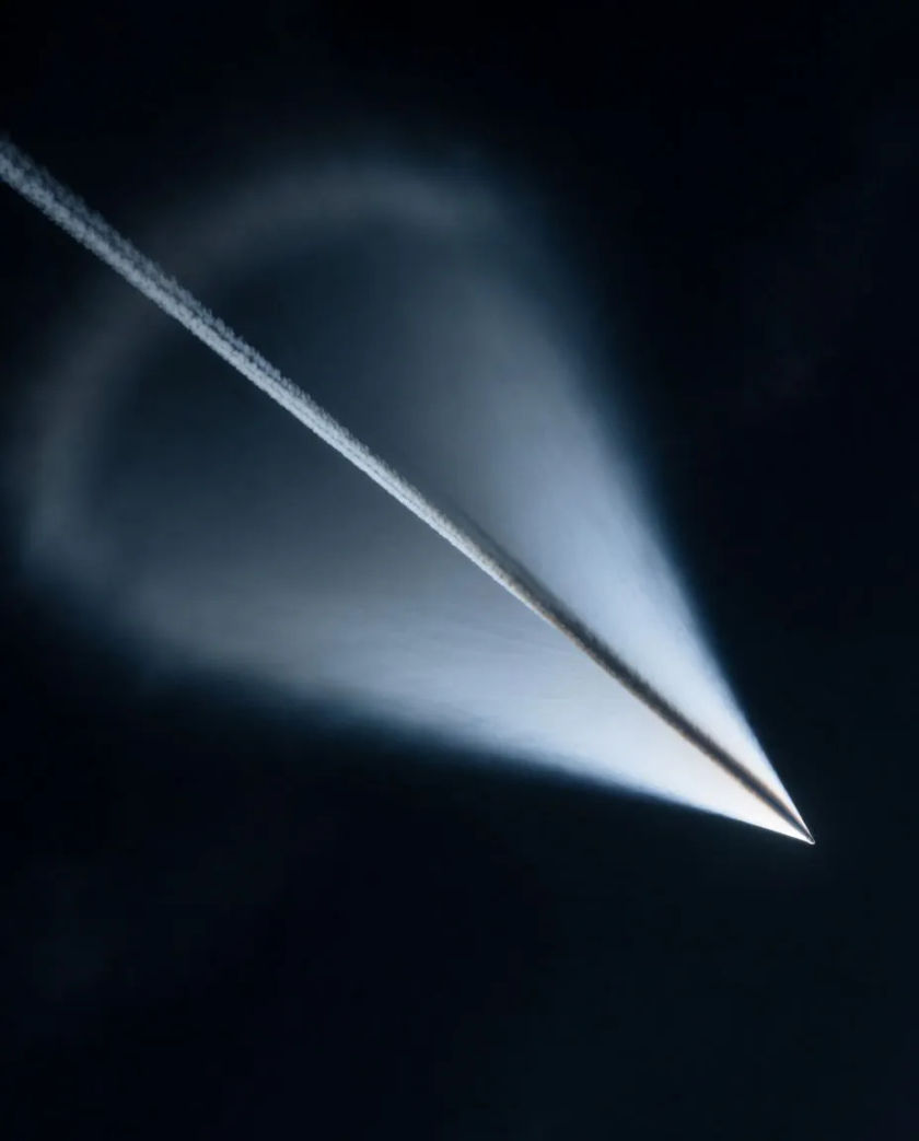A razor-thin cone of pale vapor wrapped around a dark streak of light crossing black sky, a shock ring of mist spreading behind it