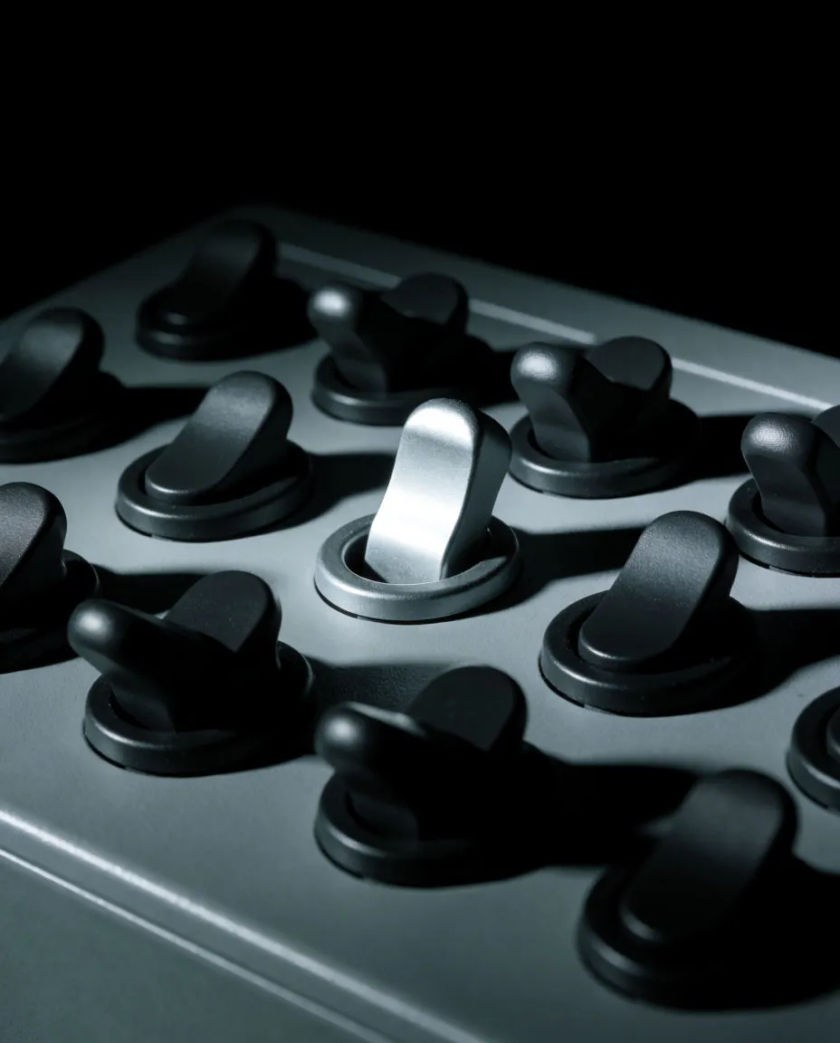 A panel of mixed switches in black and silver, one toggle flipped up and caught by a thin line of cool light, matte near-black ground