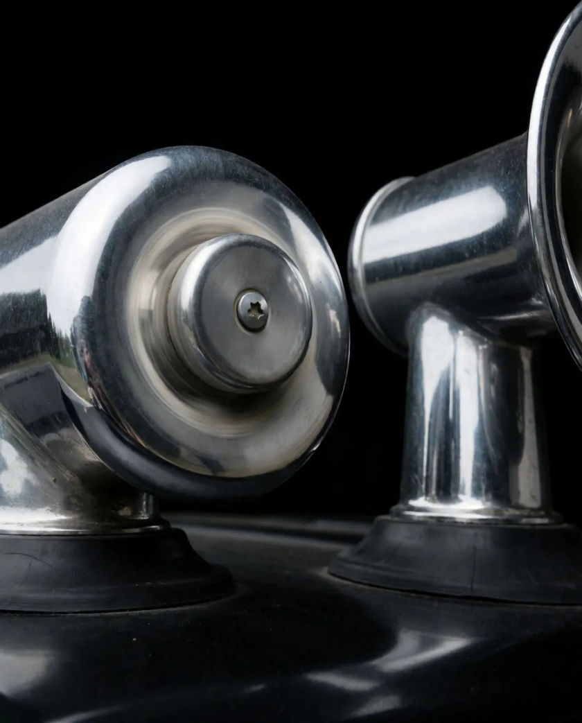 Twin chrome air horns on a cab roof with a pale shock ring of dust blown off the metal, lit hard from one side against black