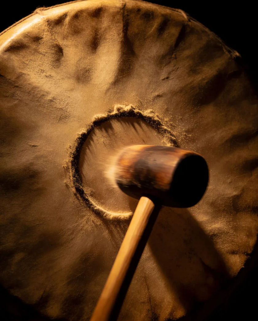 A huge stretched-hide drum struck dead centre, a ring of dust leaping off the skin in slow motion and a single shaft of amber light across it, black background