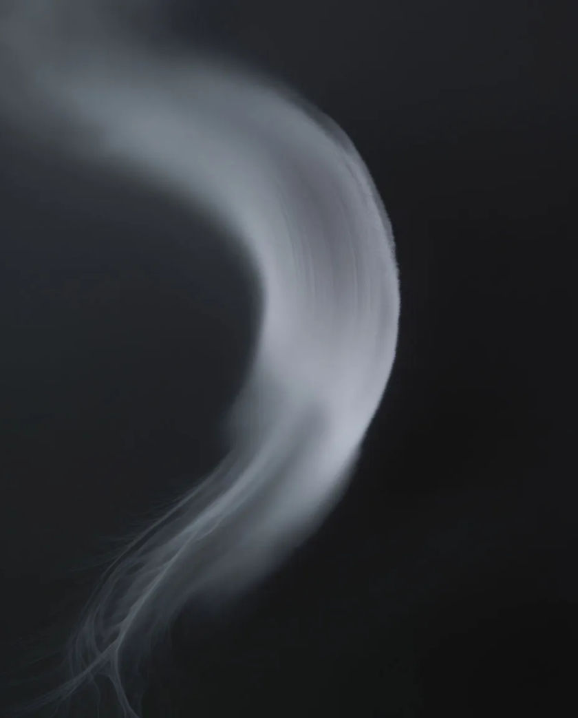 A long soft curved streak of pale mist sweeping across a dark charcoal frame, trailing into fine wisps at one end