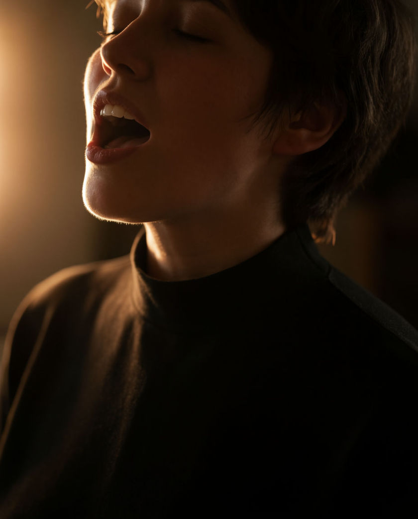 An open mouth mid-vowel in warm rim light, eyes closed, everything behind it dissolving into shadow