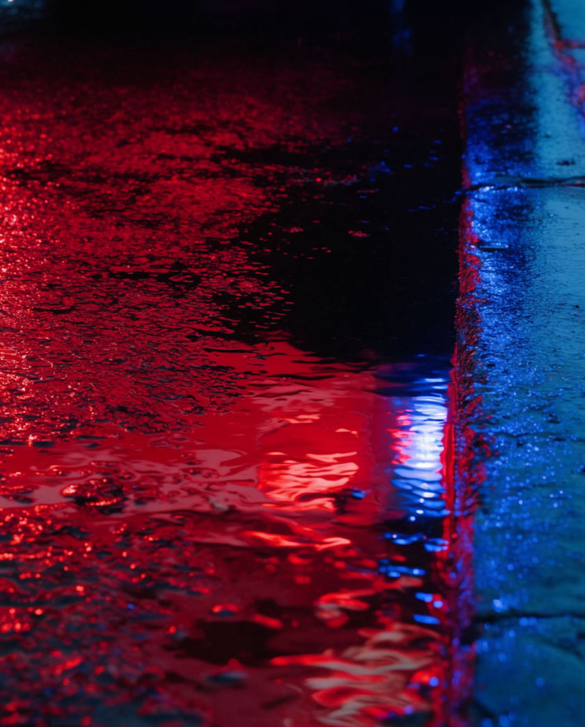 A wet street streaked with red and blue light thrown from a passing emergency vehicle, reflections filling the frame