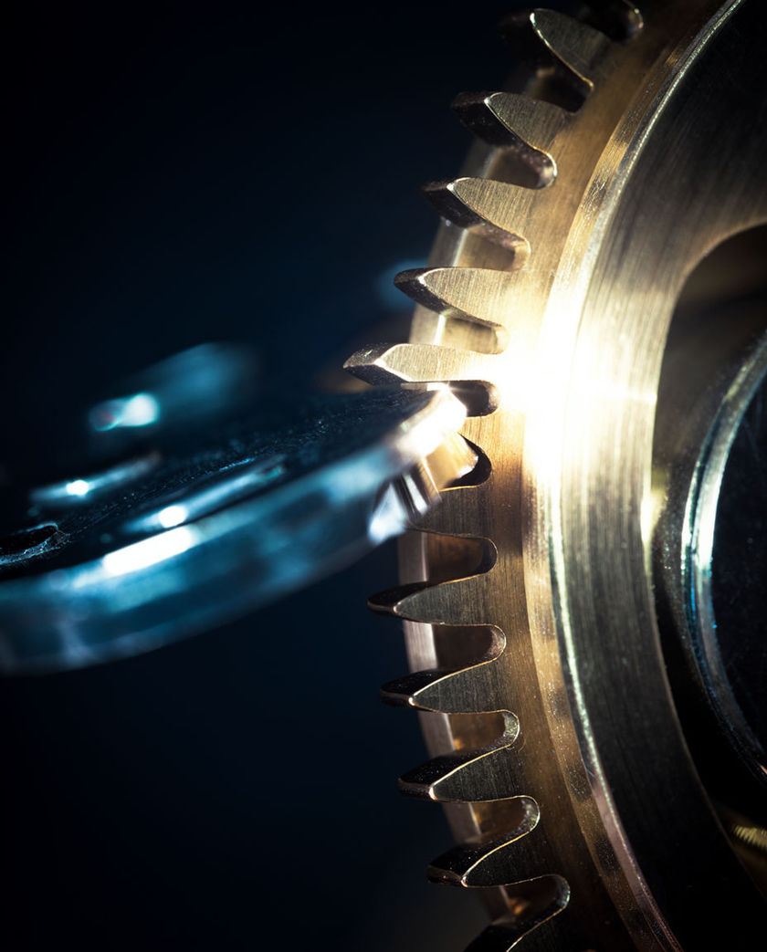 A brass escapement wheel and pallet fork in tight macro, teeth catching a single hard highlight