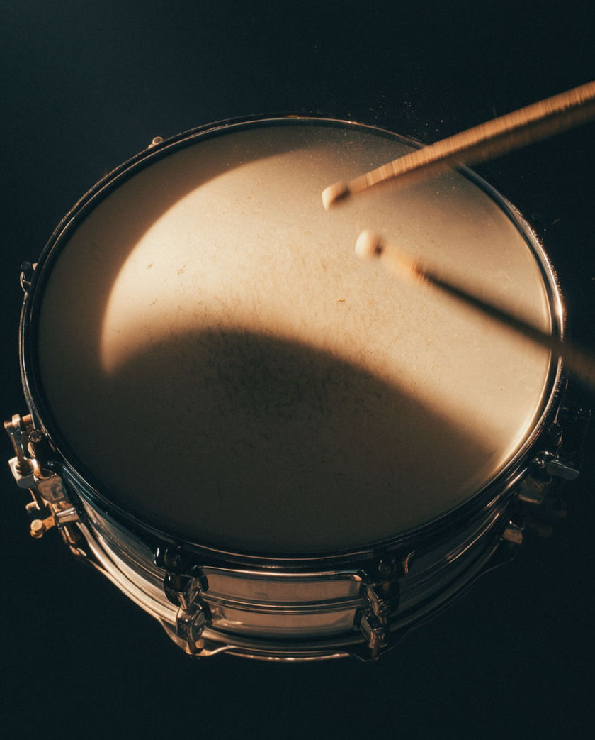 A tight snare head filling the frame from above with two stick tips blurred mid-strike and fine dust lifting off the skin