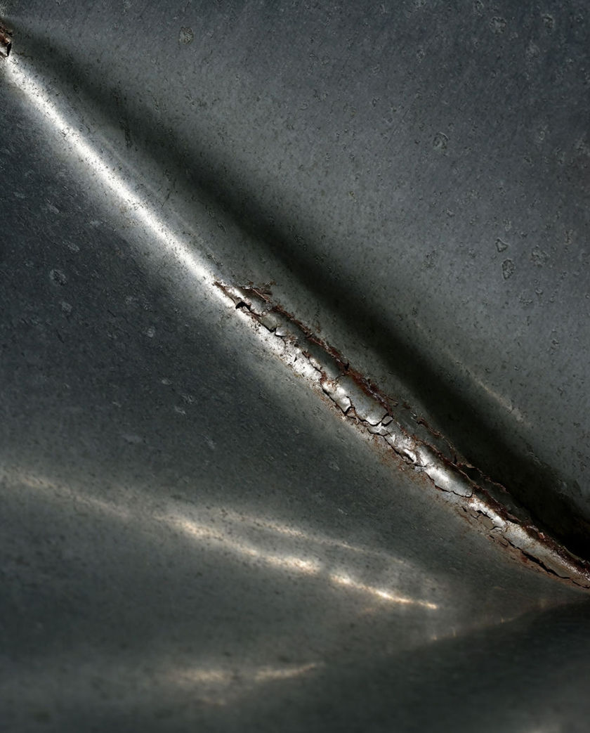 A buckled sheet of grey steel filling the frame, one cold highlight bending along a stress crease