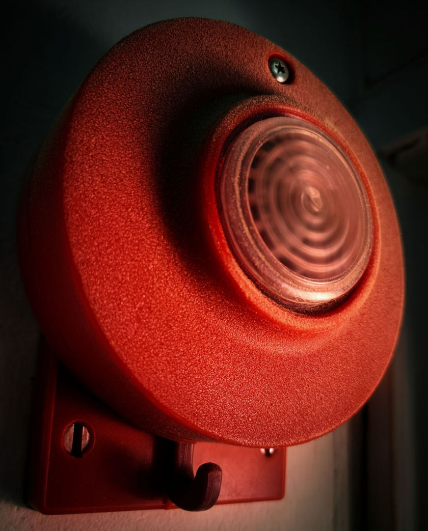 A red wall sounder shot close and slightly low, its grille catching hard red light against a dark corridor wall