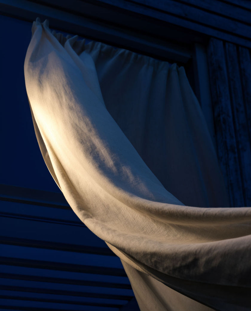 A pale curtain lifted into a slow arc by moving air, the fabric edge caught sharp against deep blue dark