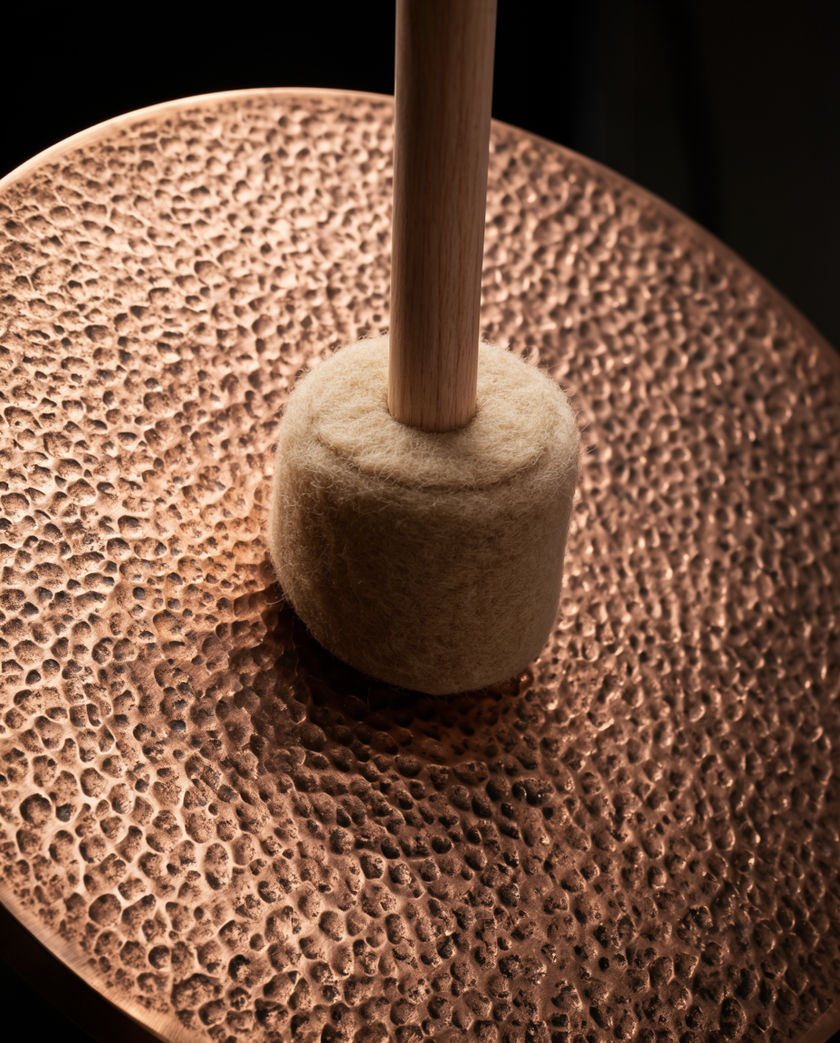 A wide hammered bronze disc lit from one side, the mallet still touching it at the moment of contact