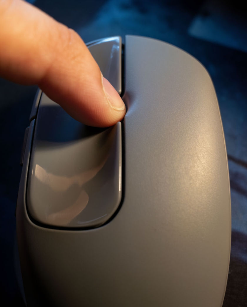 A close overhead view of a fingertip pressing a matte grey mouse button, the seam under it catching one hard edge of light