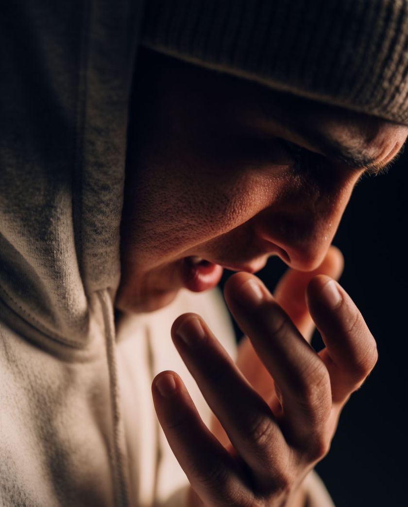 A face turned down and away in soft low light, one hand halfway to the mouth, the background falling to black