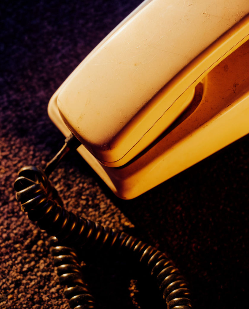 A handset lifted just clear of its cradle, cord curling below, hard light catching the worn keypad beneath it