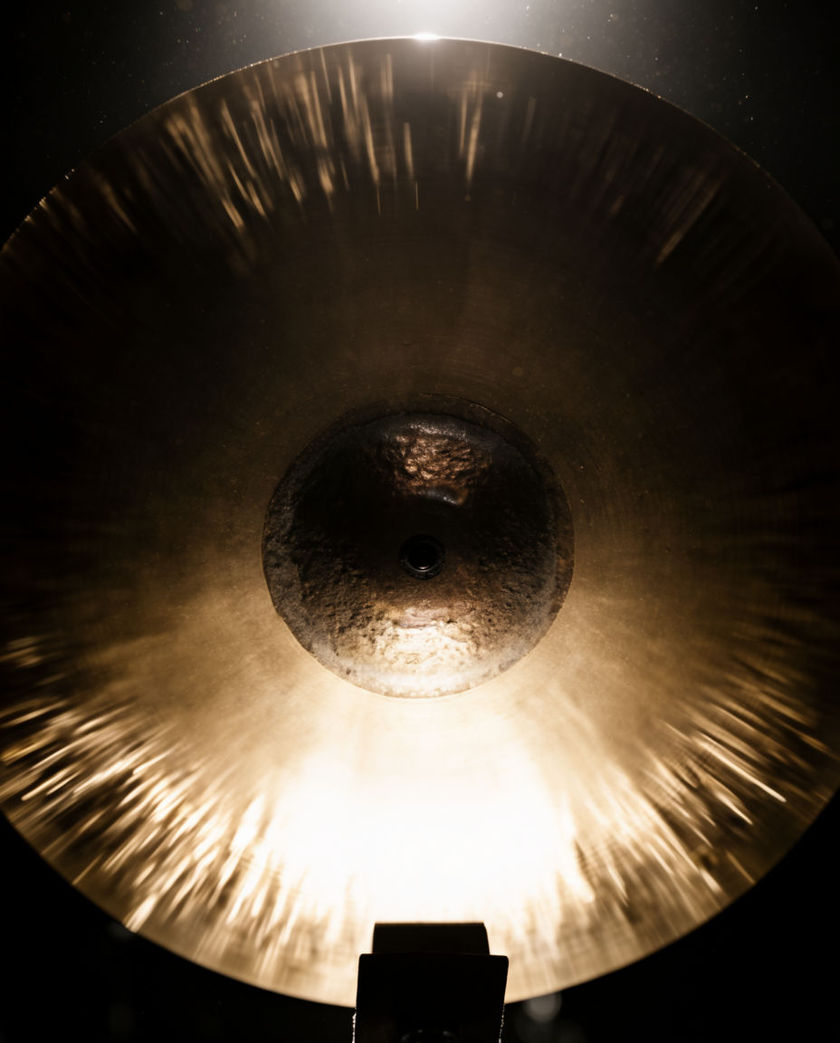 A brass cymbal shot from below with light gathering into its centre, edges dissolving into dark
