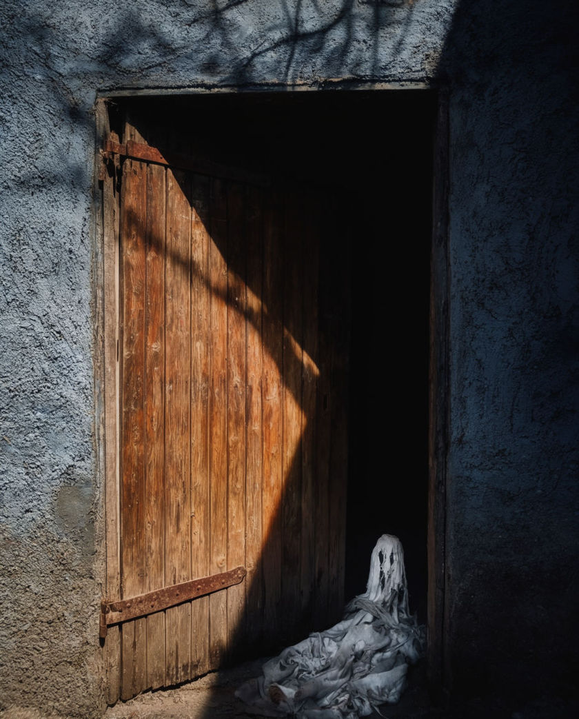 A single shaft of hard light falling across a warped wooden door left slightly ajar, the gap behind it solid black