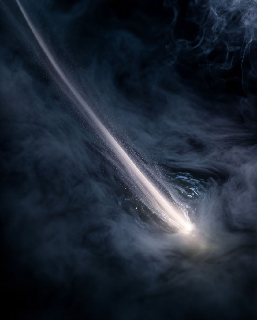 A long streak of pale light dragging through dark haze, particles pulled along its length and flaring where it ends