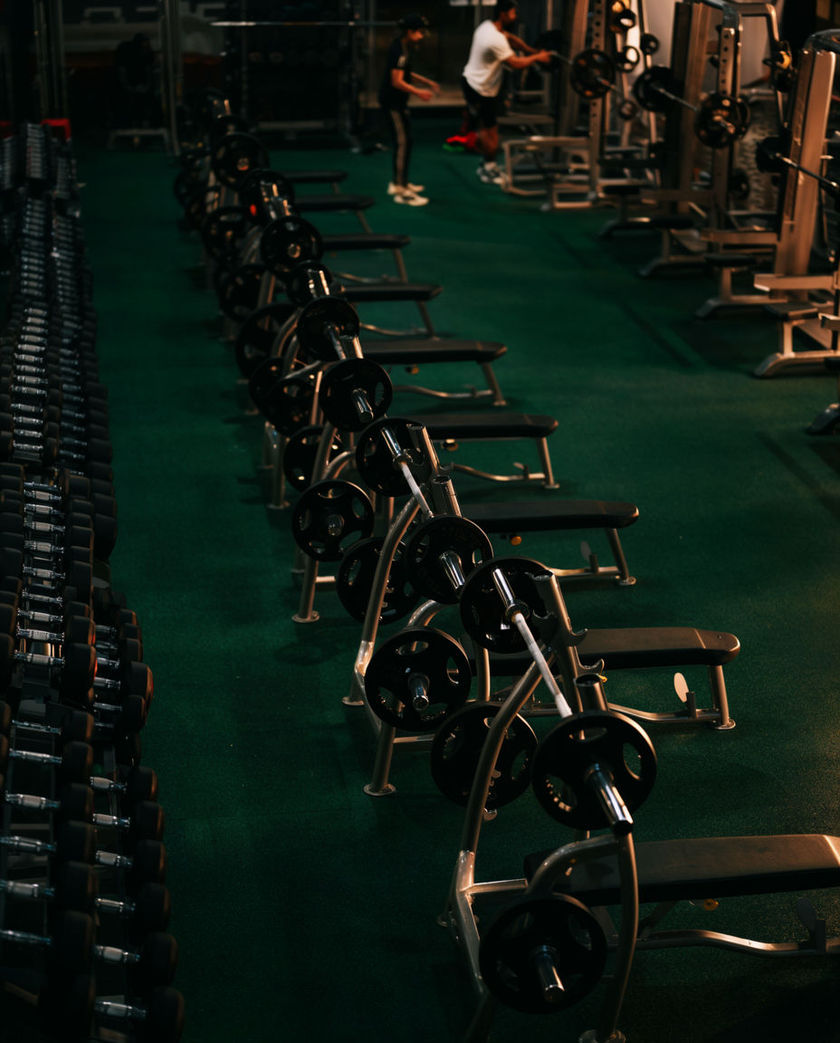 A wide gym floor lined with weight racks and treadmills under bright overhead lighting, empty of people.