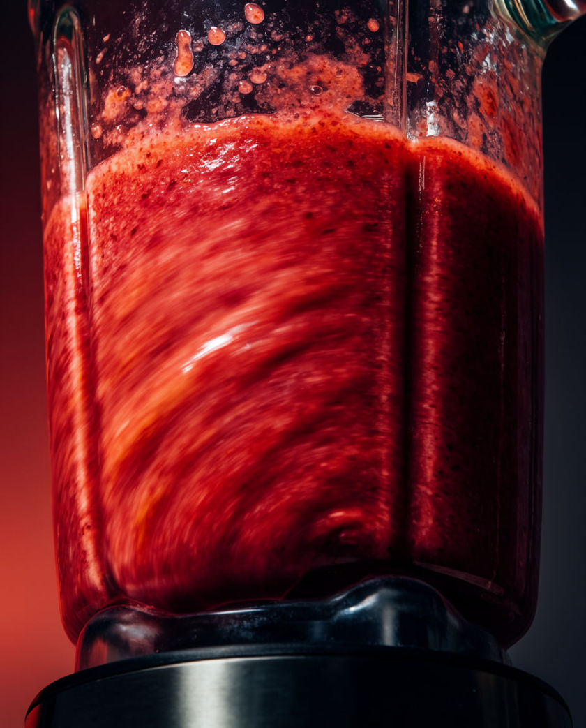 A blender jug mid-whirl with fruit blurring inside, cropped by the frame under one warm side light on a dark cool ground