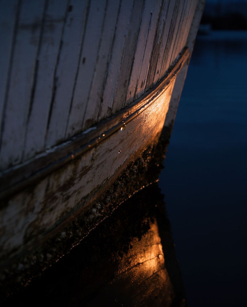 The wooden bow of a small boat cutting dark water under warm low light, spray peeling off the hull mid-motion