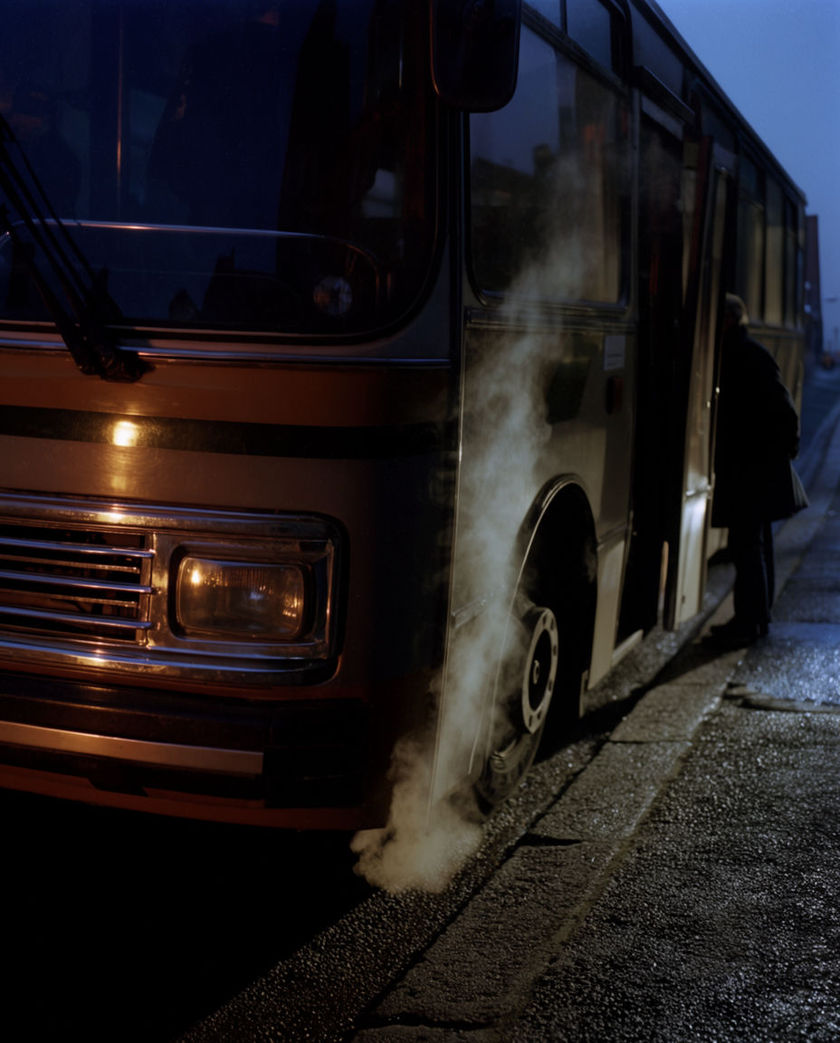 The front corner and folding door of a city bus close from a low off-axis angle under one warm light on a dark ground, exhaust haze drifting from beneath as it idles