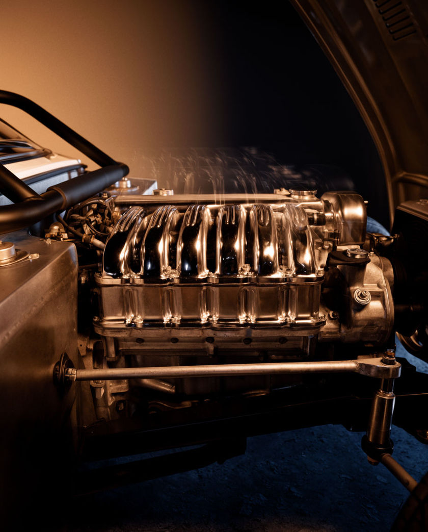 A car engine block seen close from a low off-axis angle under one warm light on a dark ground, exhaust haze rising as it runs