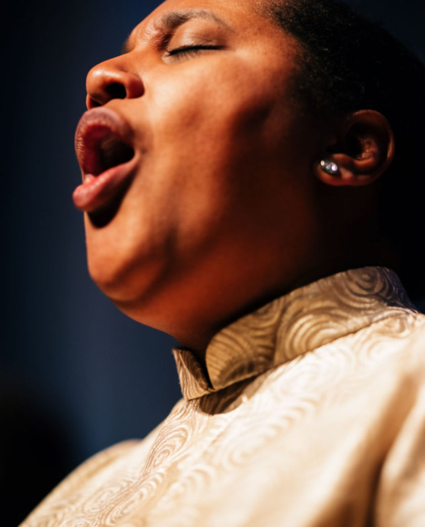 Rows of choir singers lit from one warm side against a dark ground, mouths open mid-note in a rising swell