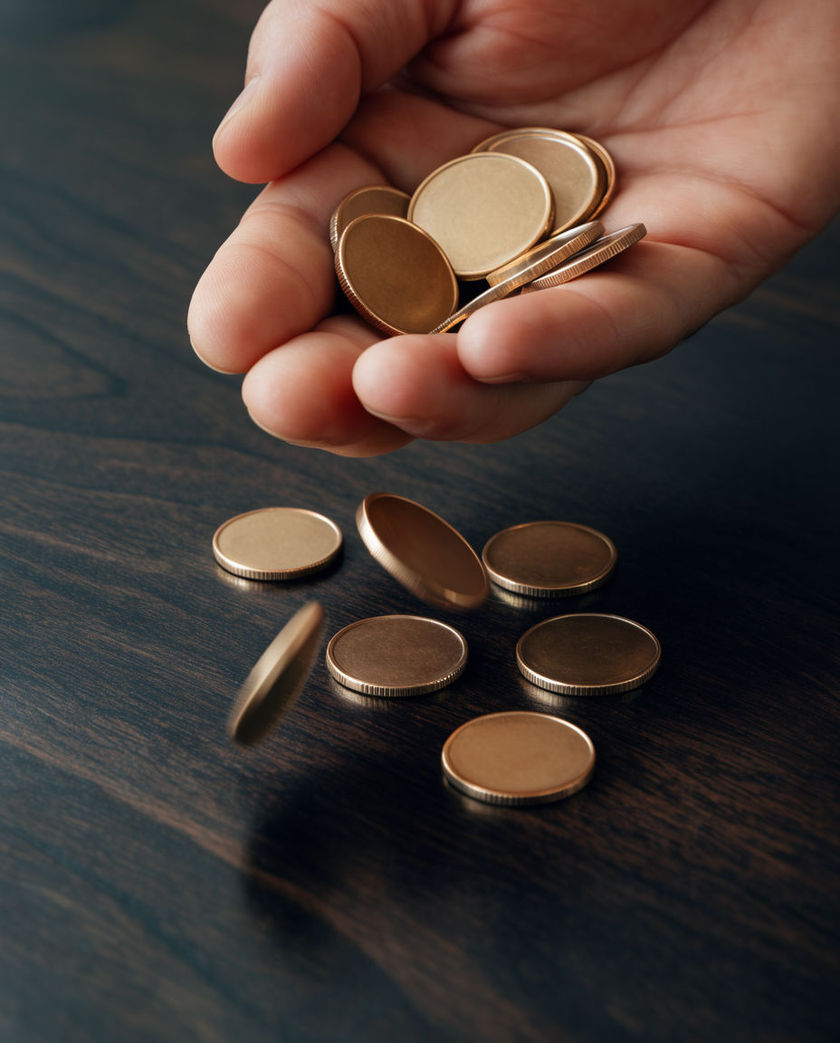 A scatter of loose coins and a brass keyring on a dark table under one warm side light, a coin mid-bounce