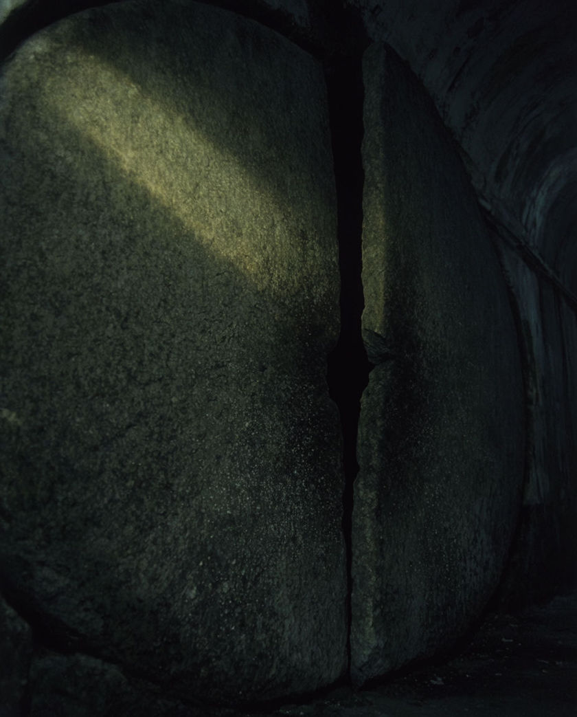 A massive stone tomb door ajar under warm low light against a cold black ground, edge caught mid-grind